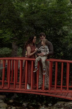 Couple sitting together on a red wooden bridge surrounded by lush green trees in a romantic garden setting.
