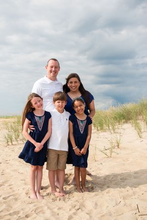 A group poses together on a sandy beach with dune grass and cloudy skies in the background.