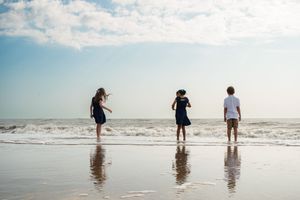 Three people stand at the edge of the ocean waves on a sunny beach with their reflections visible in the wet sand.