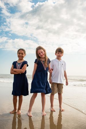 Three children standing together and smiling on a sunny beach with waves and clouds in the background.