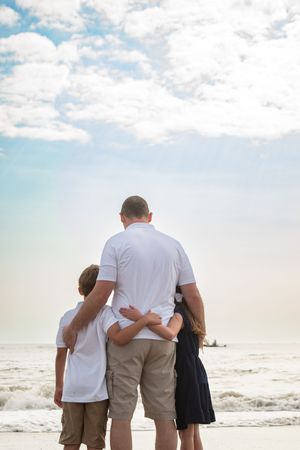 Three people stand together embracing while looking out at the ocean waves and cloudy sky at the beach.