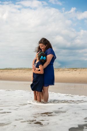 Two people in blue dresses share a tender embrace while standing in the ocean waves on a sunny beach.