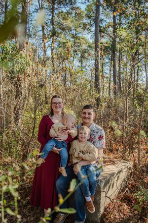 Family portrait in a wooded forest setting with autumn foliage and tall pine trees in the background.