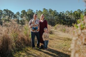 A family stands together on a nature trail surrounded by tall grass and pine trees on a sunny afternoon.