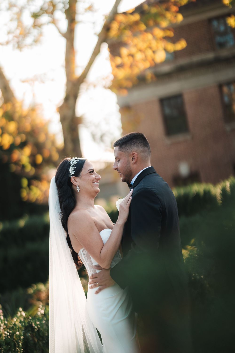 A romantic wedding couple embraces outdoors during golden hour with autumn foliage in the background.