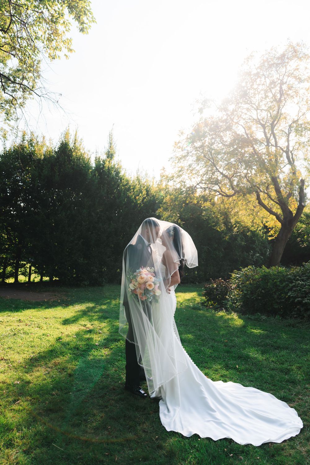 Couple in wedding attire embraces on a sunlit green lawn with trees in the background during golden hour.
