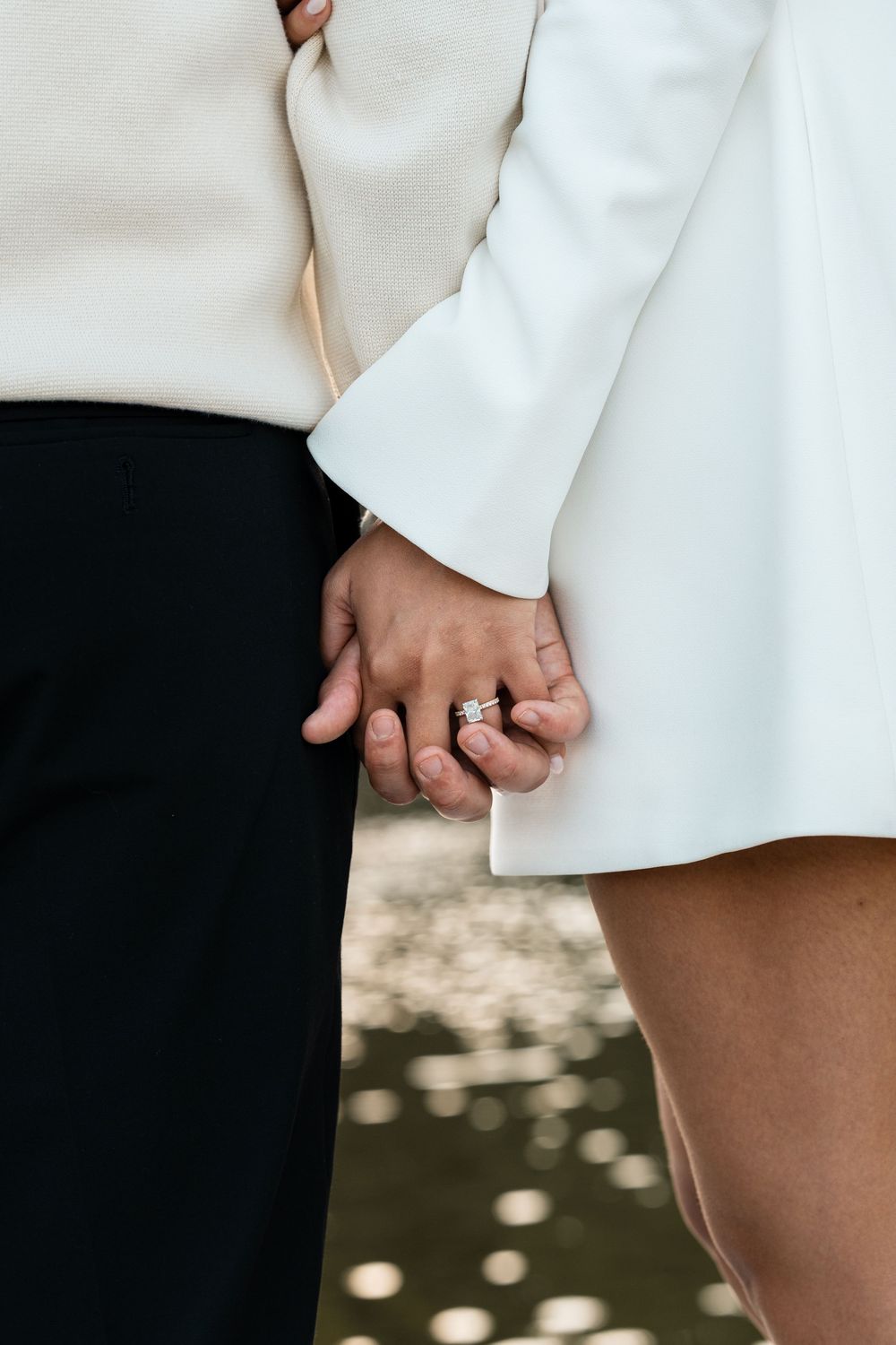 Close-up view of hands wearing an elegant diamond engagement ring against a white dress and dark background.