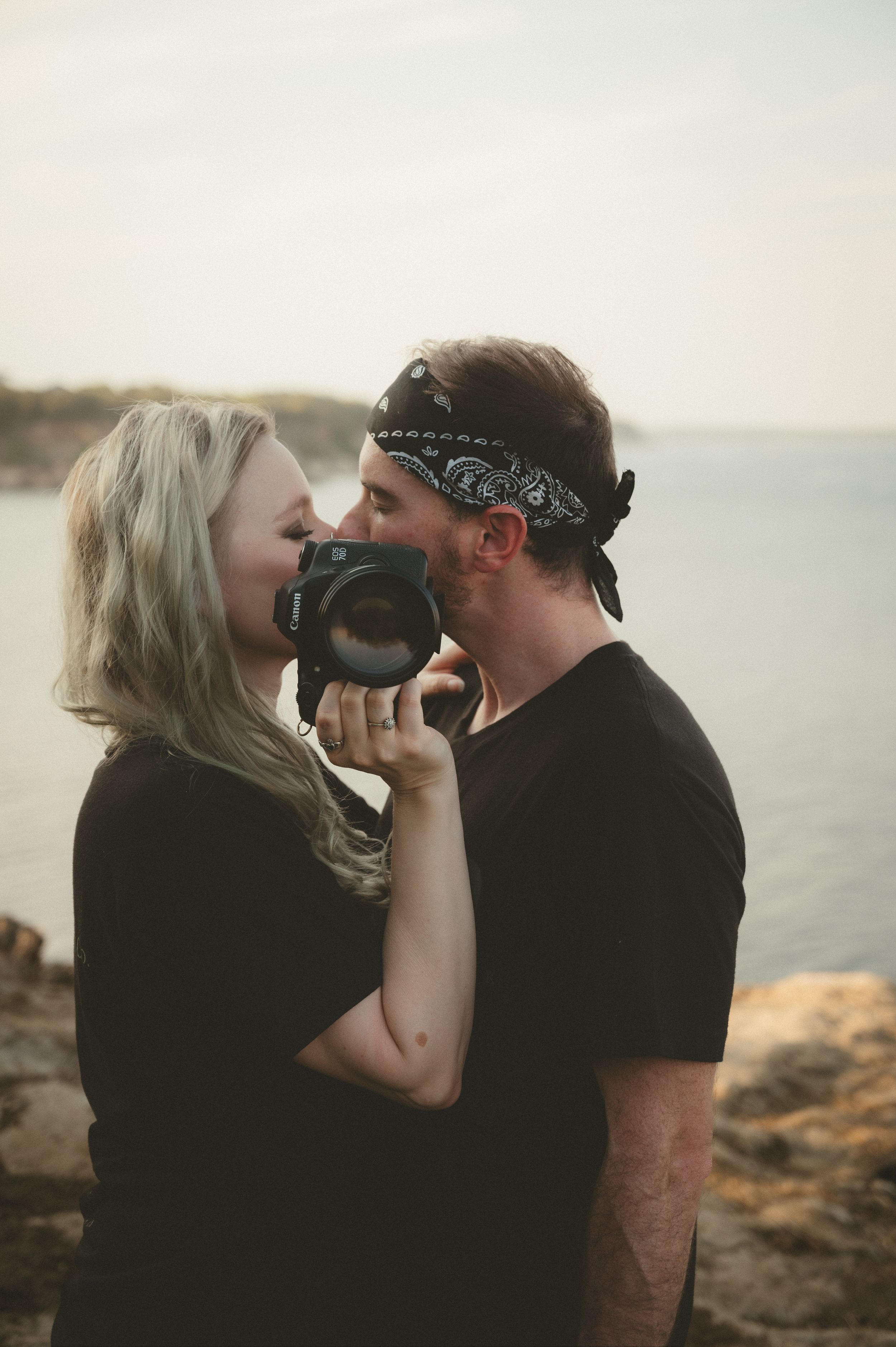 A couple shares a romantic moment while holding cameras on a rocky coastal cliff at sunset.