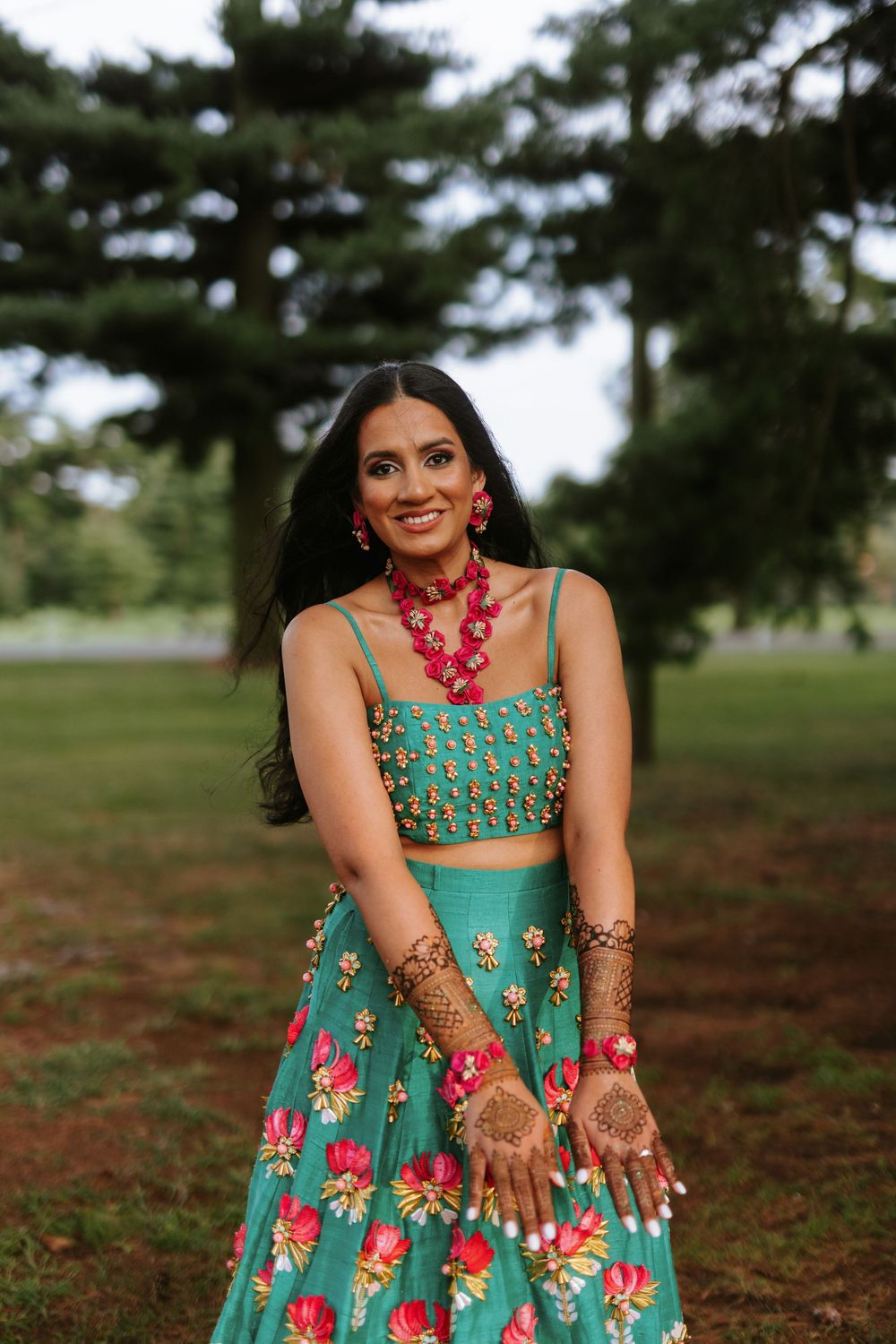 A person in a turquoise floral lehenga with red jewelry and henna designs poses outdoors near evergreen trees.