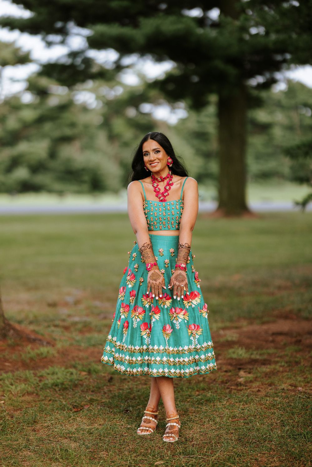 A person wearing a vibrant turquoise dress with coral floral details poses gracefully in a grassy outdoor setting.
