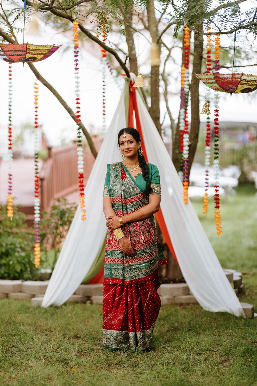 Outdoor wedding decor with white fabric drapes, red floral garlands, and lanterns hanging between trees.