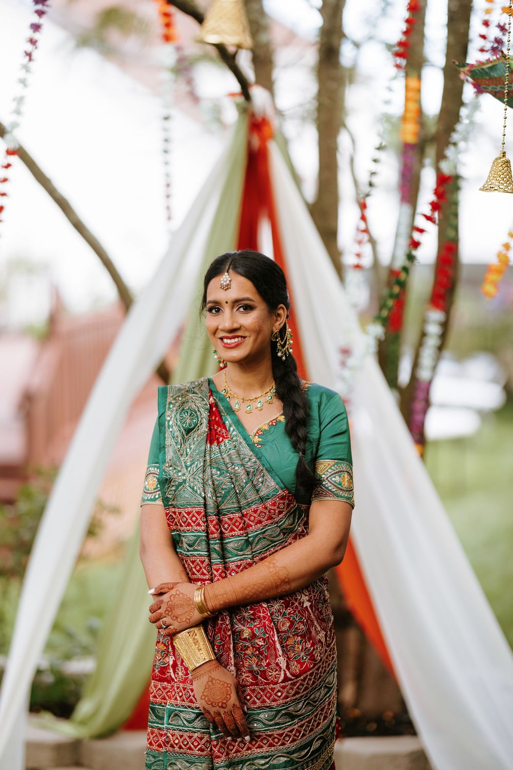 A woman in traditional South Asian bridal attire featuring a red and green saree poses elegantly against a decorated outdoor backdrop.