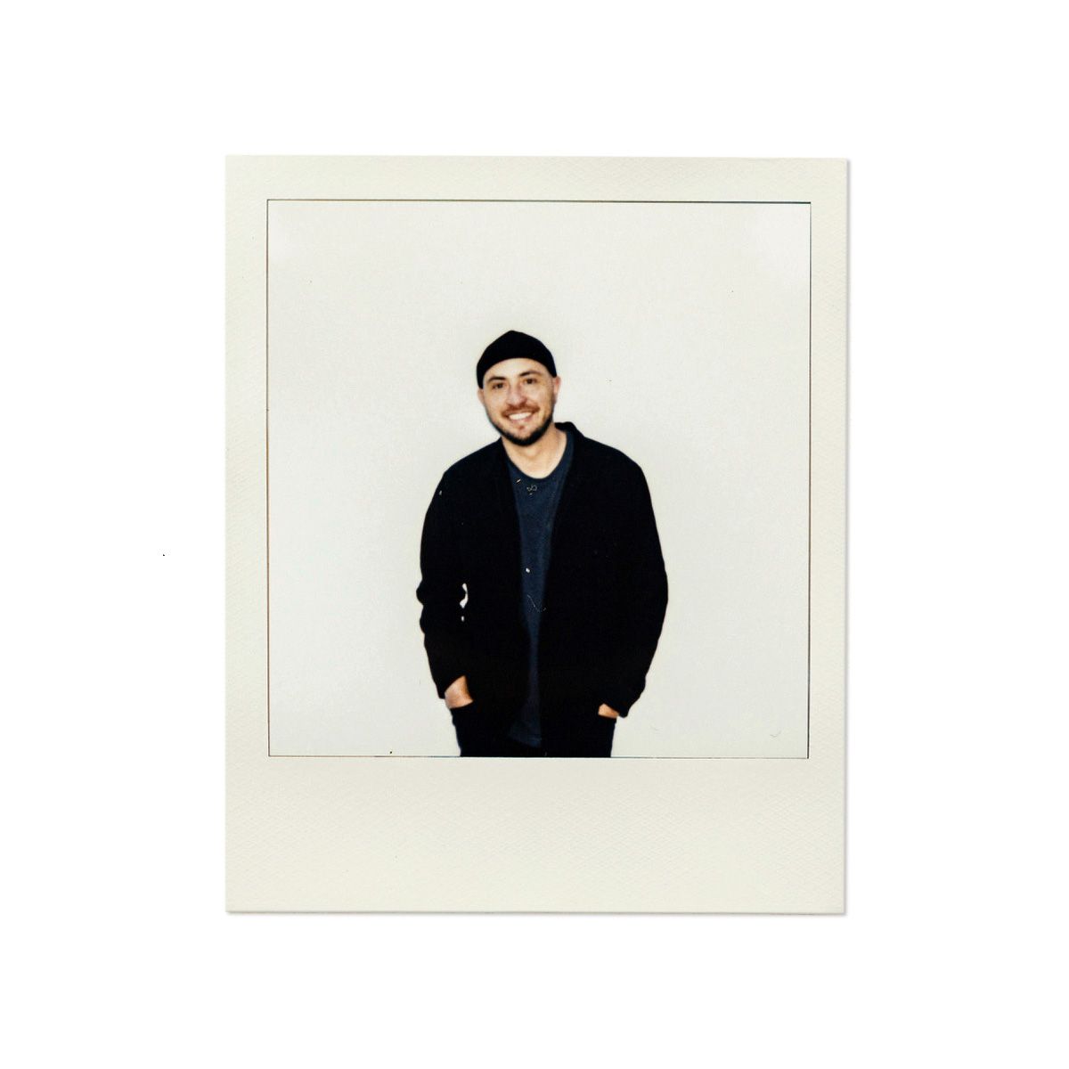 Square polaroid portrait of person in dark clothing against minimalist white background.