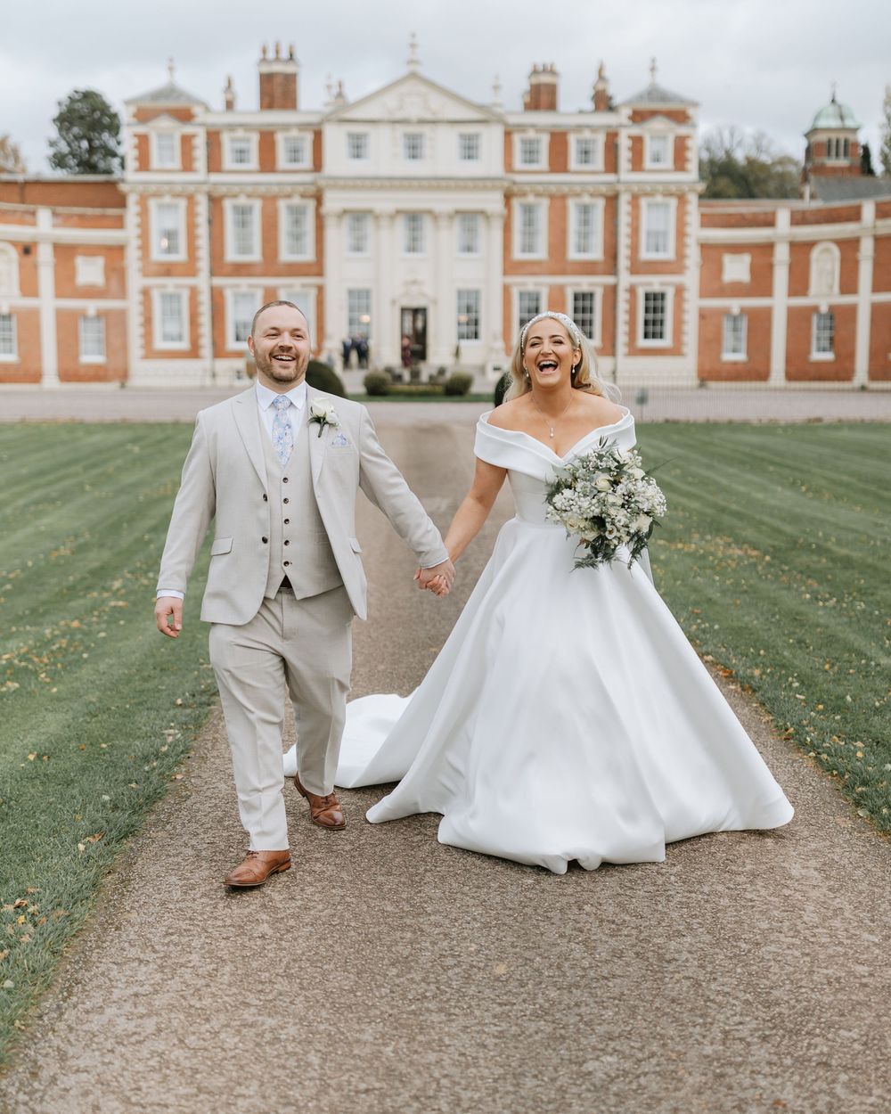 A joyful couple in wedding attire walk and twirl on a path in front of a grand historic mansion.