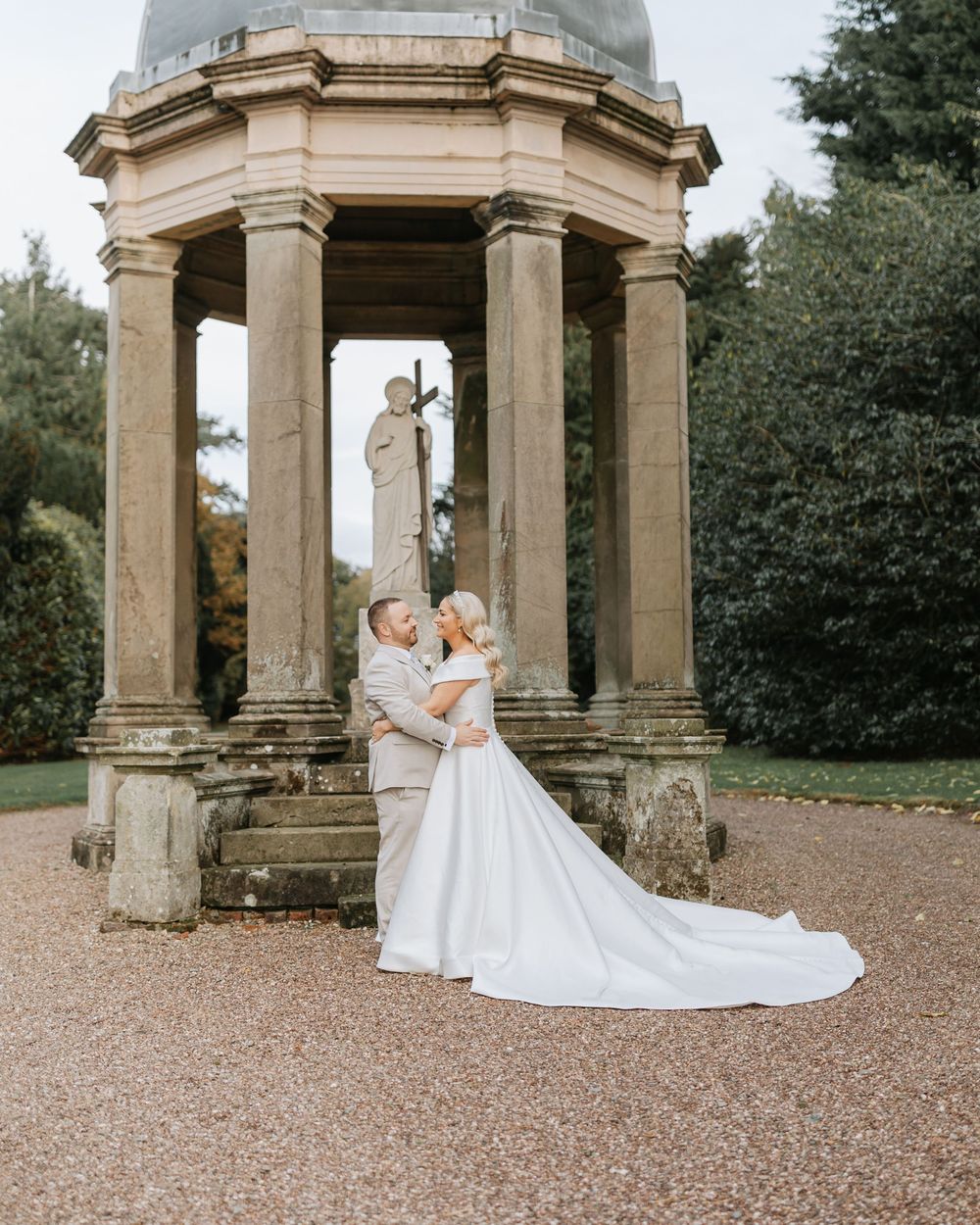 A couple in wedding attire poses together under an elegant stone gazebo in a garden setting.