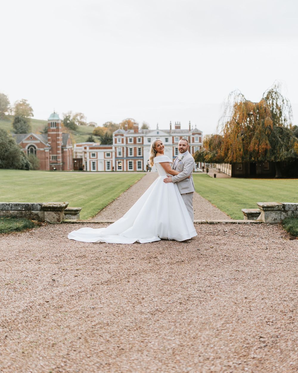 A bride in a white gown twirls on a gravel path leading to an elegant historic mansion on a sunny autumn day.