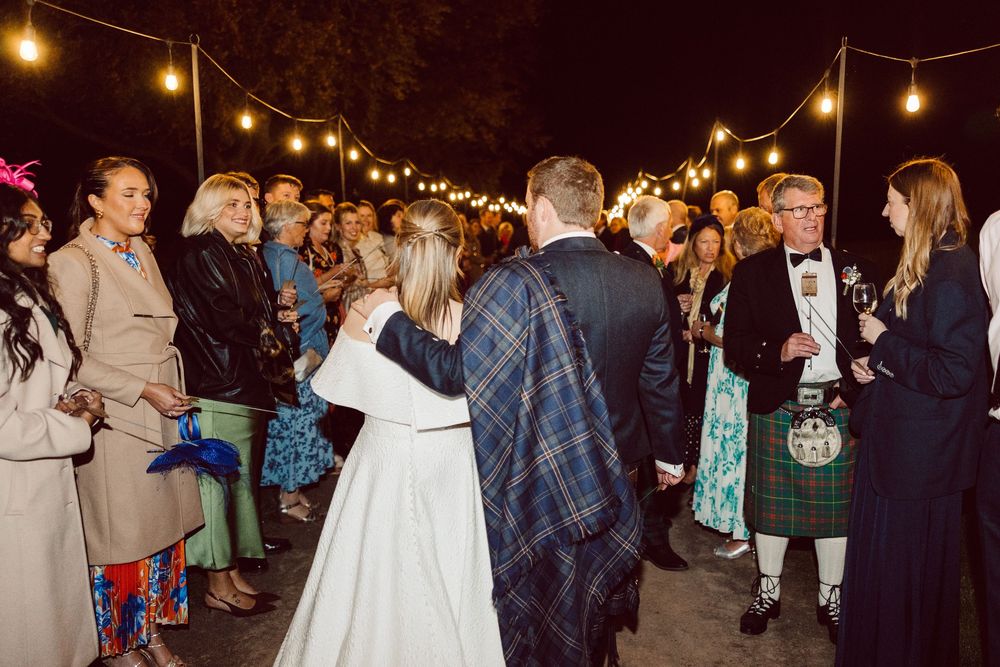 Wedding guests dance and socialize under glowing string lights at an outdoor evening reception celebration.