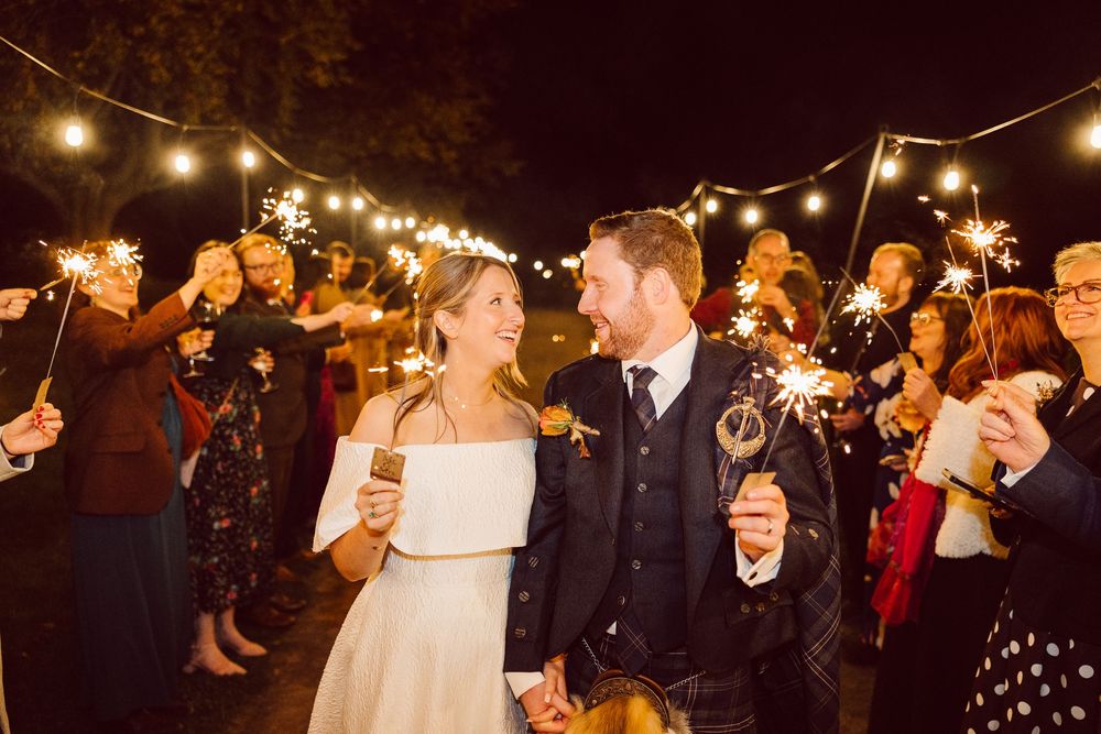 Newlyweds laugh together under twinkling lights while guests hold sparklers during evening celebration.
