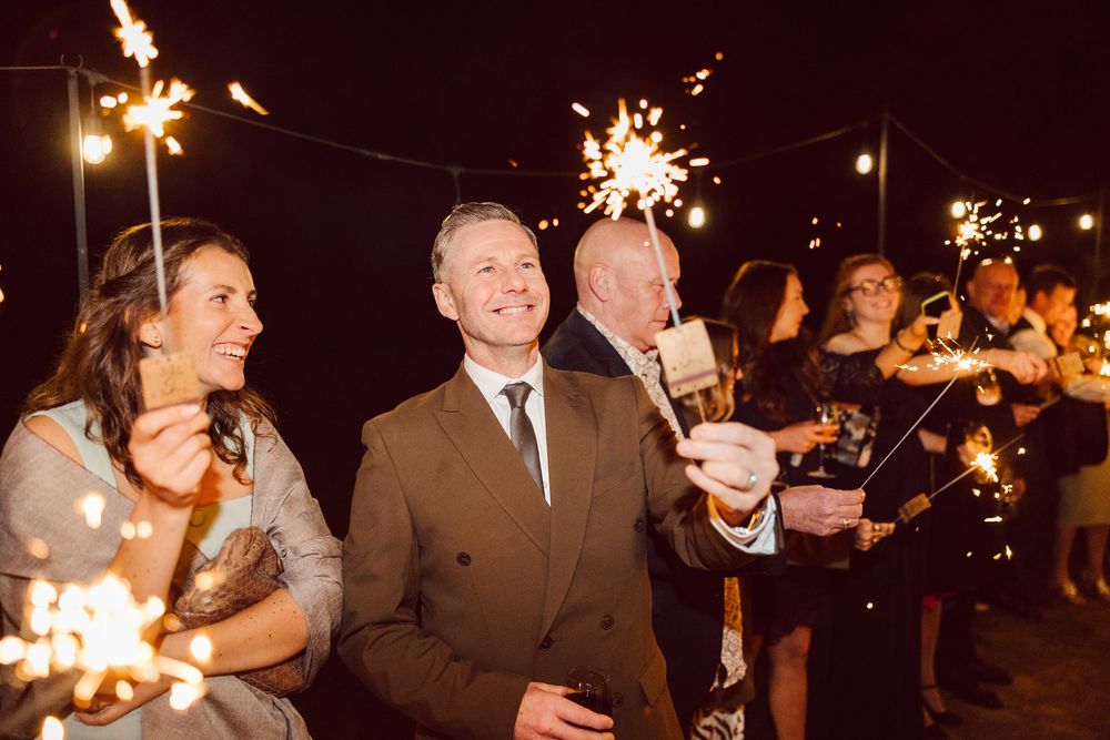 Wedding guests celebrate outdoors at night holding sparklers that illuminate the festive scene with golden light.