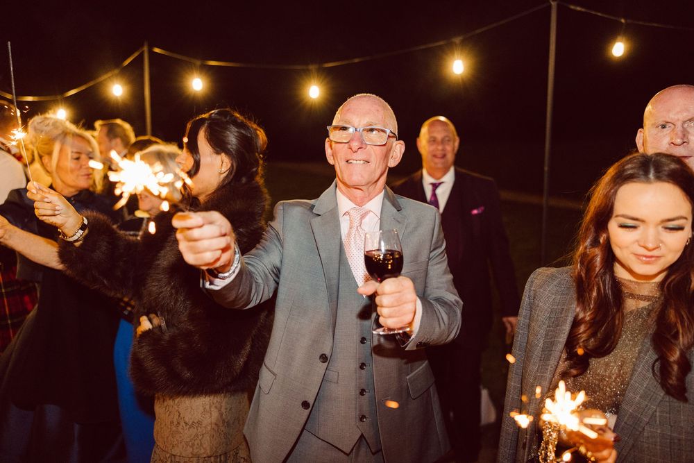 Wedding guests hold sparklers creating a magical nighttime celebration under string lights at an outdoor reception.