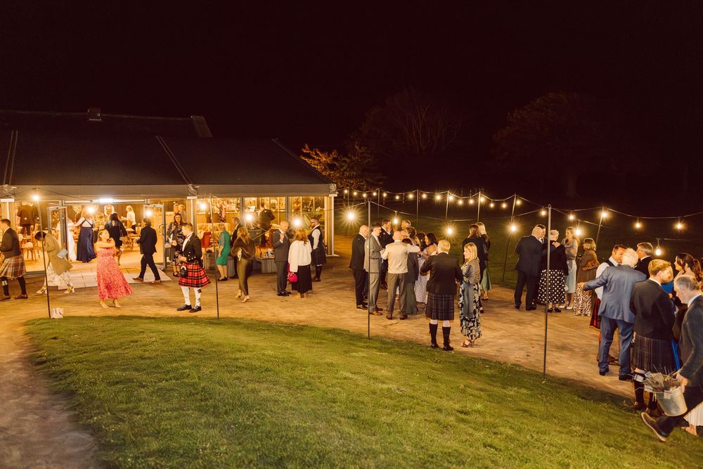 String lights illuminate a dark outdoor wedding venue with scattered guests enjoying the evening atmosphere.