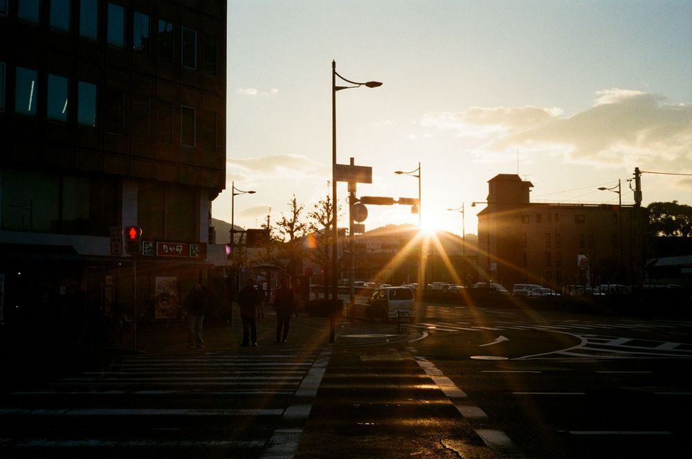 Sun setting over an urban street intersection with traffic lights and buildings silhouetted against the golden sky.