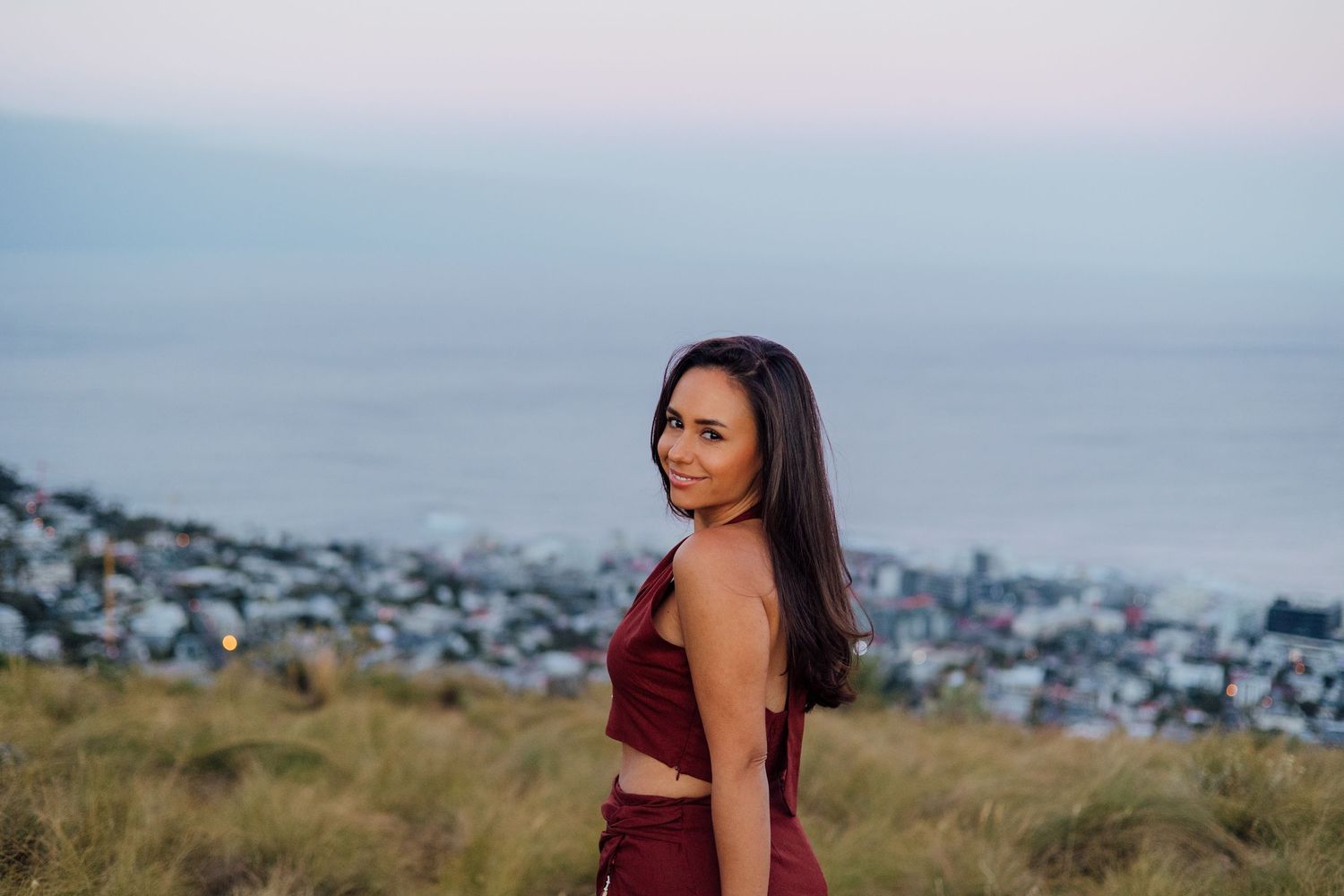 Woman in burgundy crop top poses on hillside with coastal city and ocean view at dusk.