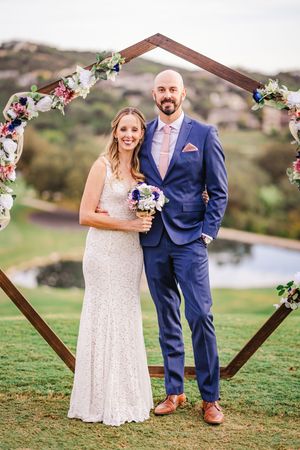 Happy couple posing together under a wood hexagon wedding arch adorned with colorful flowers outdoors.