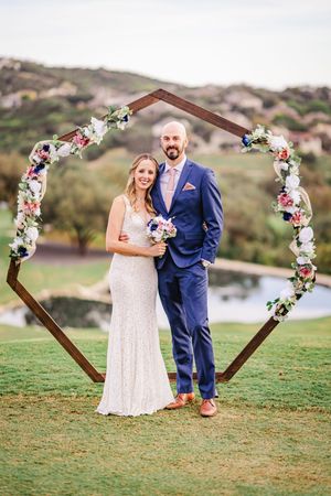 Bride and groom smiling under a hexagonal wooden floral arch on a green hillside at their outdoor wedding.