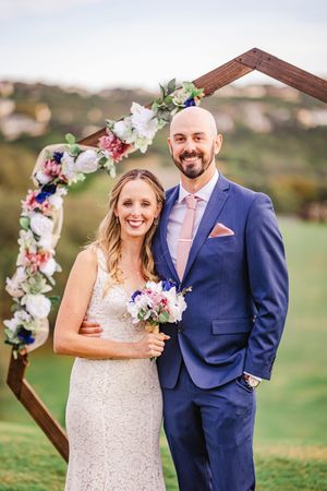 Smiling bride and groom embracing under a floral hexagonal arch at an outdoor wedding ceremony.