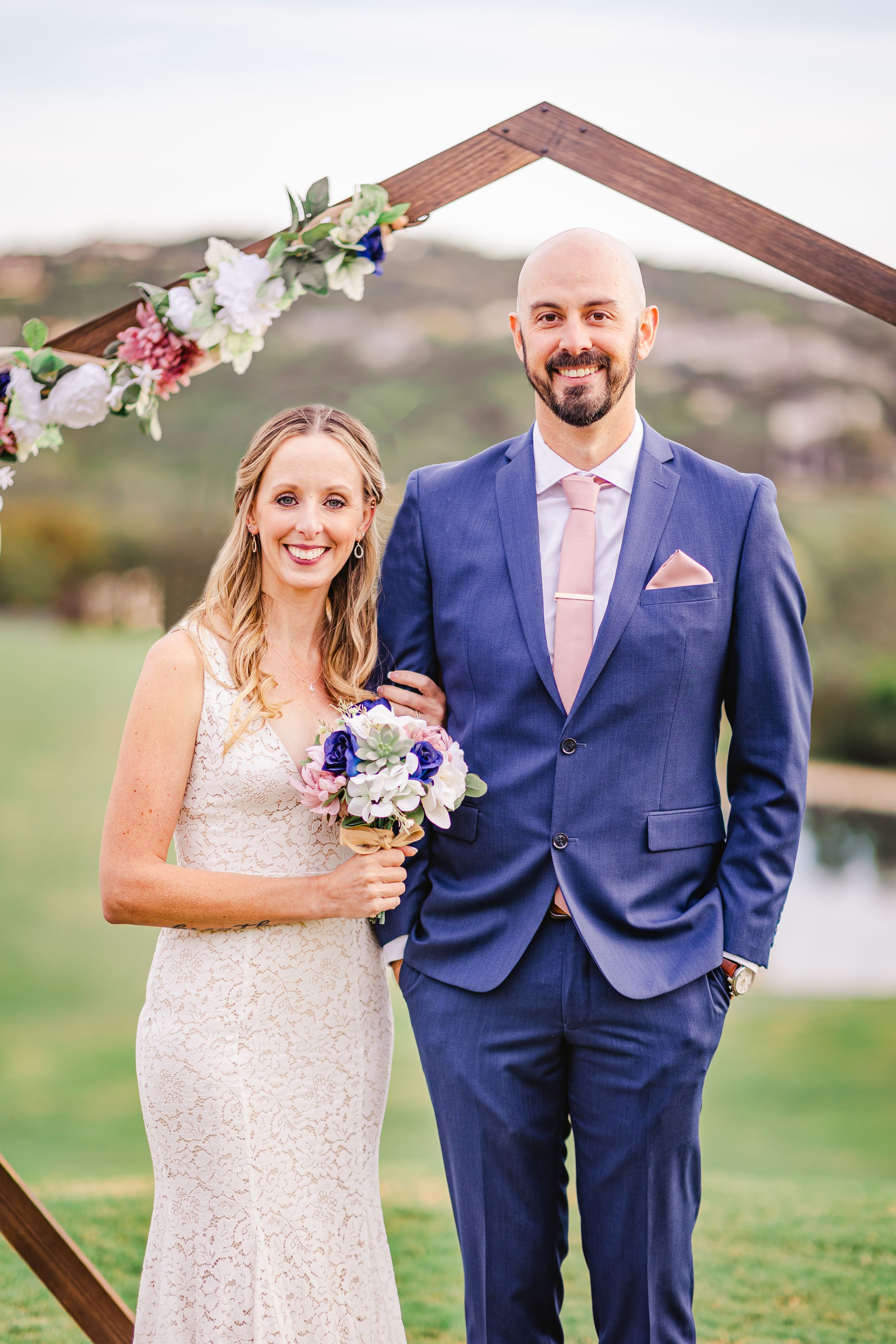 Bride holding a bouquet and groom in blue suit posing under a wooden hexagon arch at outdoor wedding.