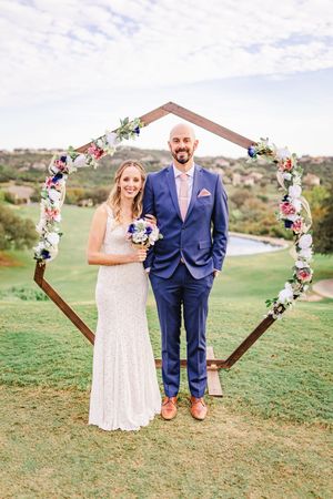 Wedding couple standing together under a large hexagonal wooden arch decorated with pink and white flowers.