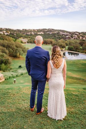 Bride and groom standing hand in hand with their backs to camera overlooking a scenic green landscape.