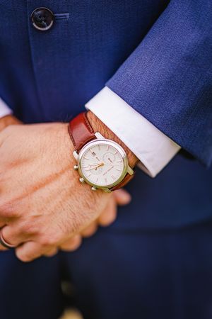 Close-up of a groom wearing a classic white-faced watch with a brown leather strap and blue suit.