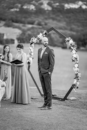Black and white photo of groom standing at the altar under a hexagonal floral arch during wedding ceremony.
