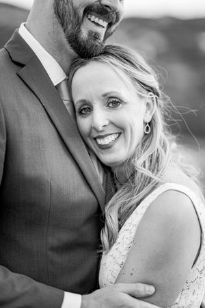 Black and white photo of smiling bride leaning against groom in suit at outdoor wedding.
