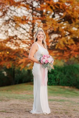 Bride in white gown holding pink bouquet with fall foliage background at outdoor wedding.