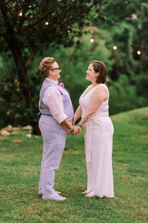 Two brides holding hands in garden wedding ceremony with string lights in background.