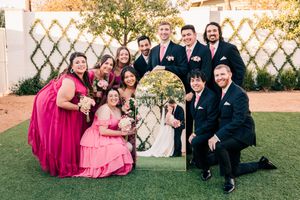 Wedding party posing with decorative arch, bridesmaids in pink gowns, groomsmen in navy suits.