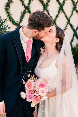 Bride and groom kissing, groom in navy suit with pink tie, bride holding pink rose bouquet.
