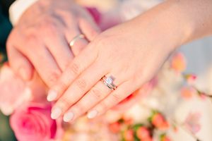 Close-up of bride and groom hands showing diamond engagement ring and wedding band over flowers.