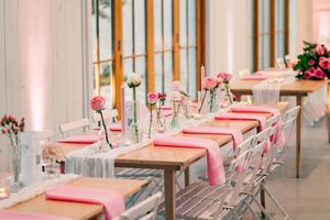 Wedding reception tables with pink napkins, bud vases with pink roses, and white chairs.