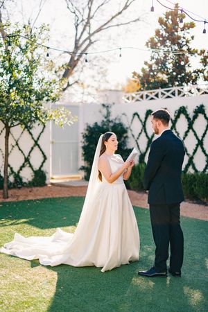 Bride in white gown reading vows to groom on green lawn at outdoor venue.