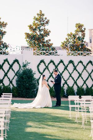 Bride and groom exchanging vows on a green lawn with white lattice wall backdrop.