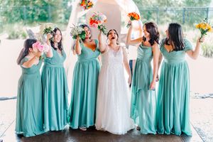 Bride in white lace gown celebrates with five bridesmaids in teal dresses holding colorful bouquets.
