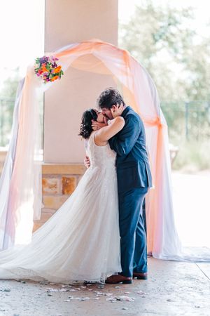 Bride and groom share first kiss under elegant peach draped arch with colorful floral arrangement.