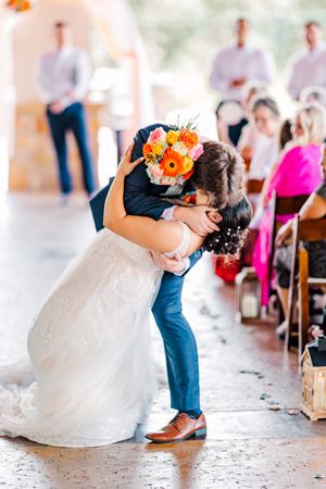 Groom dips bride in white sparkle gown for romantic kiss during wedding ceremony recessional.