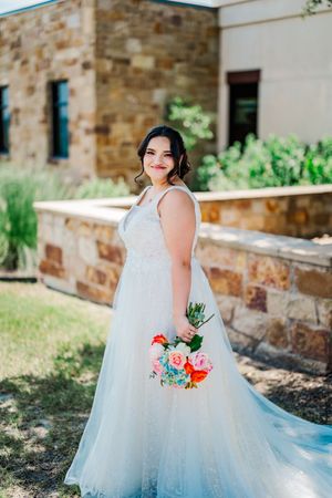 Radiant bride in white A-line wedding gown poses outdoors holding colorful bouquet near stone building.