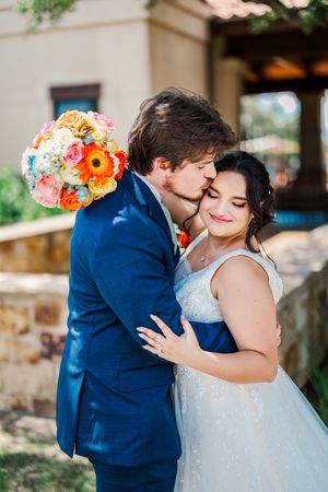 Groom in navy suit kisses bride on cheek while embracing her outdoors at wedding venue.