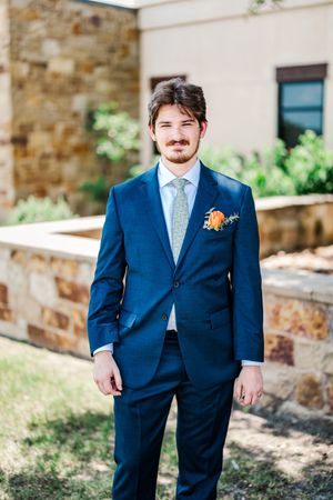 Handsome groom in navy blue suit with floral tie and orange boutonniere poses outdoors.